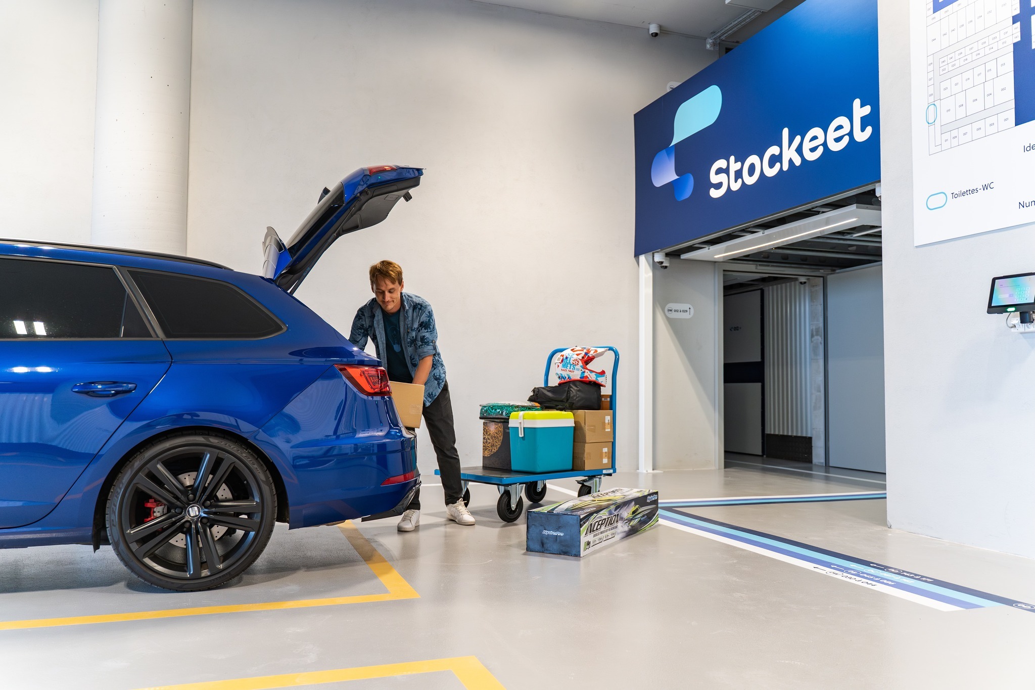 Un homme décharge un carton d'un coffre de voiture bleue sur un chariot dans un centre de self-stockage Stockeet pour un déménagement réussi.